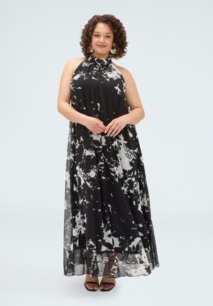 Kvinde med krøllet hår iført sort-hvid mønstret ærmeløs halter maxi-kjole og sorte åbne tå-sandaler, står mod en ensfarvet baggrund.