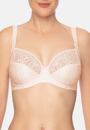 Femme portant un soutien-gorge rose clair avec des détails en dentelle fleurie et des bretelles réglables, montrée des épaules à la taille sur un fond uni.