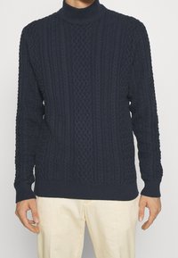 Maglione navy lavorato a maglia con collo alto. Presenta trecce strutturate, polsini e orlo a coste. Realizzato in un tessuto spesso e caldo.
