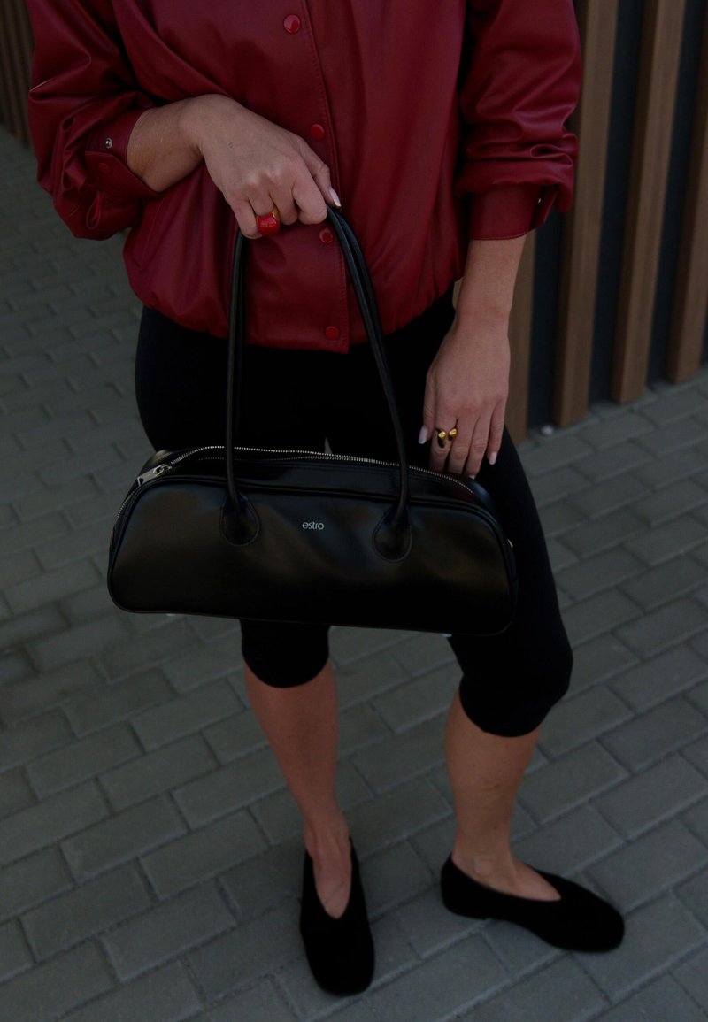 Černá kožená taška s zipovým uzávěrem, kulatým tvarem a dvojitými uchy. Nosená s burgundským sako a černými legínami.