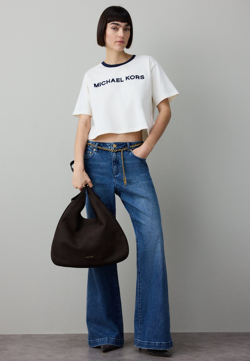 Witte cropped t-shirt met marineblauwe afwerking en "MICHAEL KORS" logo, gecombineerd met wijde blauwe spijkerbroek en een zwarte hobo-tas.