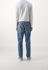 Jasnoniebieskie dżinsy z denimu o prostym kroju, w pięciokieszeniowym stylu, z naszywką z tyłu. Noszone z białą koszulą z długim rękawem i szarymi trampkami.