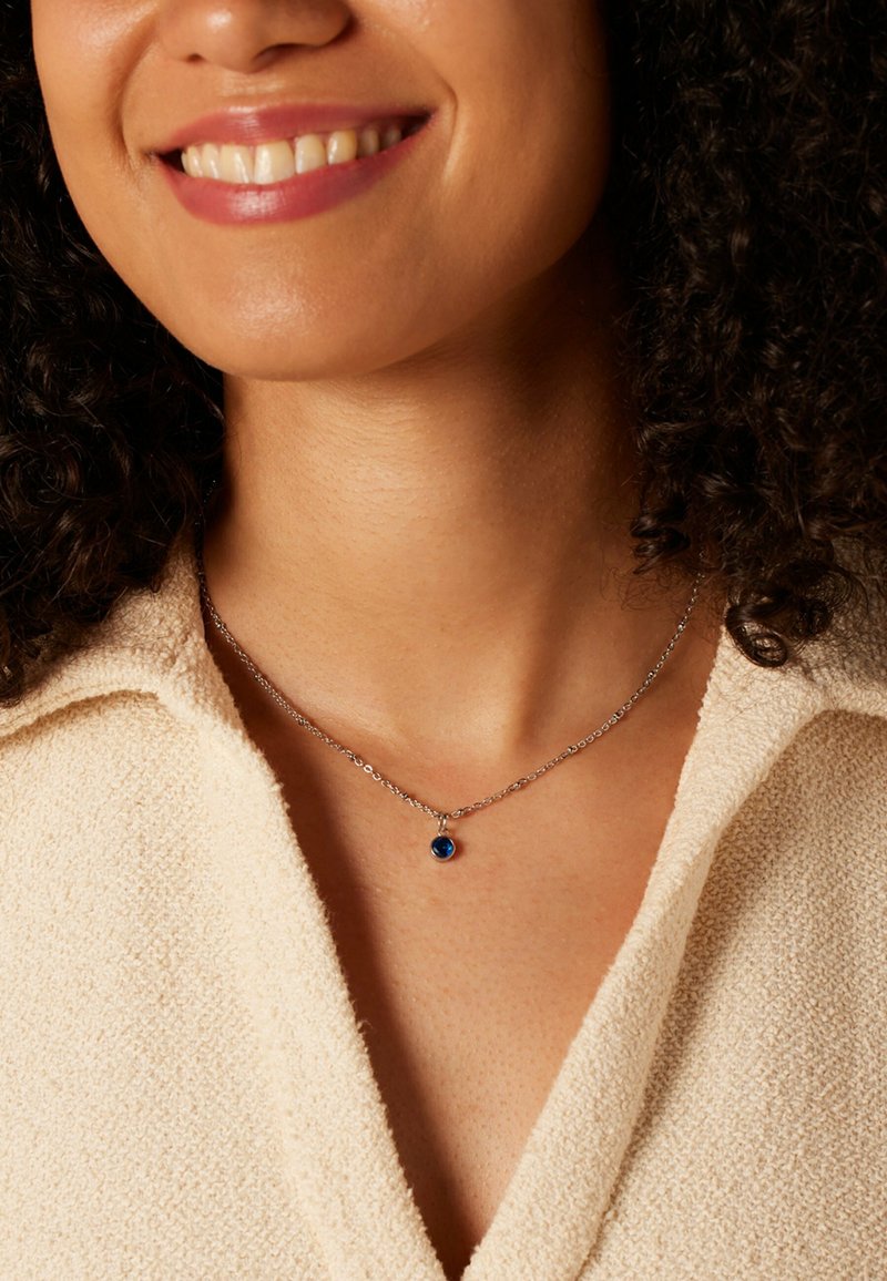 Zilveren ketting met een ronde, blauwe edelsteenhanger, tegen een gewatteerde beige stoffen achtergrond.