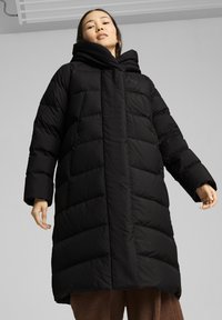 Manteau d'hiver noir avec un col haut, texture matelassée et longues manches larges. Présente une coupe droite atteignant le mollet.