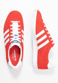 Röda mockaskor för träning med vita läderdetaljer, som har tre vita ränder, vita snören och en strukturerad gummisula.