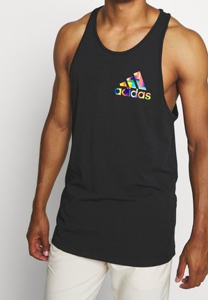 Muskelbepackte Person trägt ein schwarzes ärmelloses Tanktop mit farbigem Adidas-Logo auf der Brust, kombiniert mit hellen Hosen.