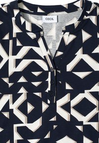Top a rayas en azul marino y crema con formas geométricas, que cuenta con un tapeta con tres botones negros y un escote redondeado. Fabricado con un tejido suave.