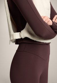 Een bruine longsleeve top, gelaagd met een witte fleece vest. Het vest heeft een gestructureerd oppervlak en een zijsluiting met een gesp. Straksluitende leggings.