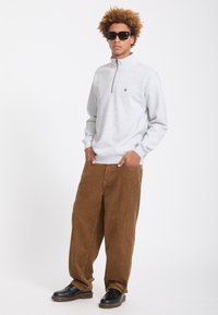 Jeune homme portant des lunettes de soleil noires, un sweat-shirt gris clair à fermeture éclair quart, un pantalon en velours côtelé marron et des chaussures noires, debout les mains dans les poches.
