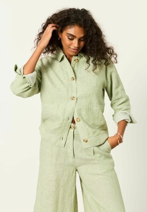 Žena s kučeravými vlasmi, oblečená v svetlozelenom ľanovom saku na gombíky a zodpovedajúcich širokých nohaviciach, jedna ruka v vrecku, druhá si dotýka vlasov.