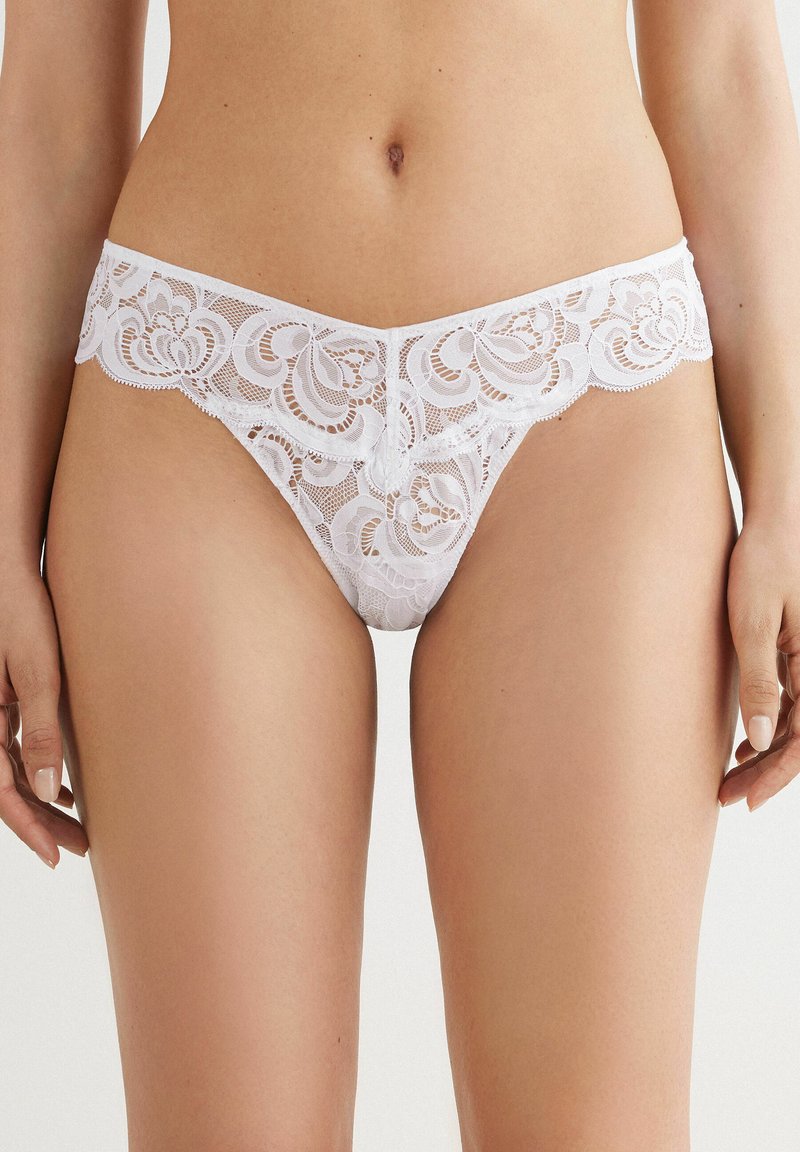 Thong en dentelle blanche avec des motifs floraux, des bords ondulés et un délicat détail en ruban au centre avant. Texture douce et transparente.