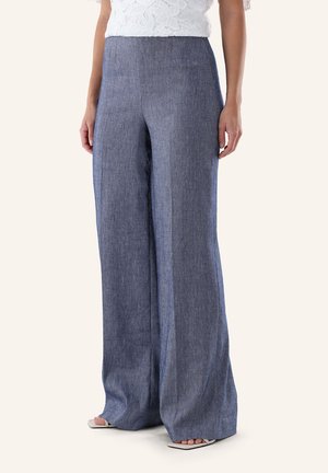 Frau trägt hoch taillierte, weit geschnittene blaue Leinenhose mit weißem Spitzentop und weißen Sandalen mit quadratischer Spitze, steht vor einem einfarbigen Hintergrund.