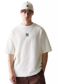 Giovane uomo che indossa un berretto beige e marrone dei New York Yankees, una maglietta bianca strutturata con il logo verde NY e pantaloni marroni, in piedi con una mano in tasca.