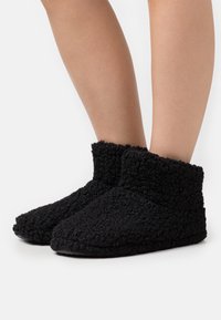 Black, fluffy slipper made of textured fabric. Ankle height with a soft finish and rounded toe design. Non-slip sole.