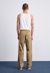 Tan cargo pants made of sturdy fabric with a relaxed fit, featuring dual back pockets and a woven logo patch. Worn with a white tank top.