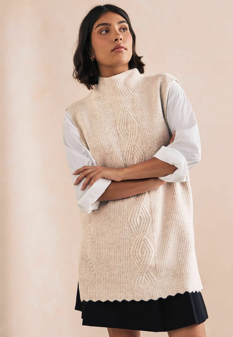 Maglione senza maniche lavorato a maglia di un leggero beige, con un colletto alto, un texture a coste e un motivo a diamante. Indossato sopra una camicia bianca con le maniche arrotolate.