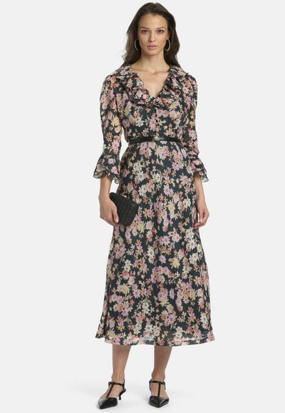 Femme portant une robe midi noire à motifs floraux avec col et manches à volants, ceinture noire, sac à main noir, boucles d'oreilles créoles et chaussures noires pointues.