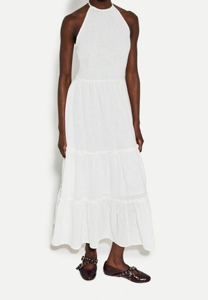 Persona che indossa un vestito maxi bianco senza maniche a balze e scarpe piatte marrone scuro con cinturini e occhielli in metallo.