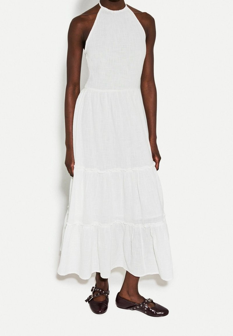 Personne portant une robe maxi blanche sans manches à volants et des chaussures plates marron foncé avec des brides et des œillets en métal.