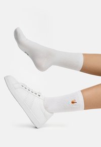 Calcetines de algodón blancos con textura acanalada, que presentan un pequeño icono de bebida naranja. Combinados con zapatillas blancas de corte bajo.