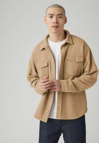 Lysebrun bomuldsskjorte med knapper, to forlommer, krave og løs pasform, båret over en hvid t-shirt og mørke jeans.