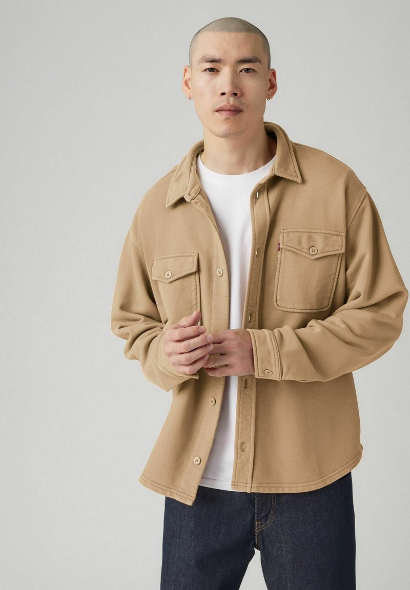 Beige katoenen overhemd met knopen, twee zakken aan de voorkant, een kraag en een losse pasvorm, gedragen over een wit T-shirt en donkere spijkerbroek.