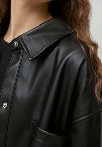 Giacca camicia in pelle nera con un ampio colletto, chiusura frontale a scatto e tasca laterale. Presenta una texture liscia e una finitura lucida.