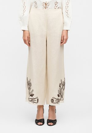 Kvinde iført beige højtaljede linnedvide bukser med sort blomstret broderi nær kanterne og sorte åbne tå-hæle.