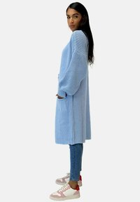 Cardigan lungo aperto in maglia azzurro chiaro, con ampie maniche morbide e tasche frontali. Abbinato a jeans aderenti e sneakers.