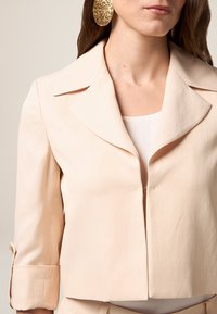 Jachetă scurtă de culoare bej deschis, cu un guler structurat, manșete cu butoane și o textură netedă. Prezintă un închidere frontală cu fermoar.