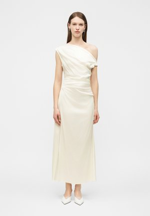 Kvinde står iført en gulvlængde, off-white satin kjole med den ene skulder blotlagt og hvide spidse sko foran en ensfarvet baggrund.