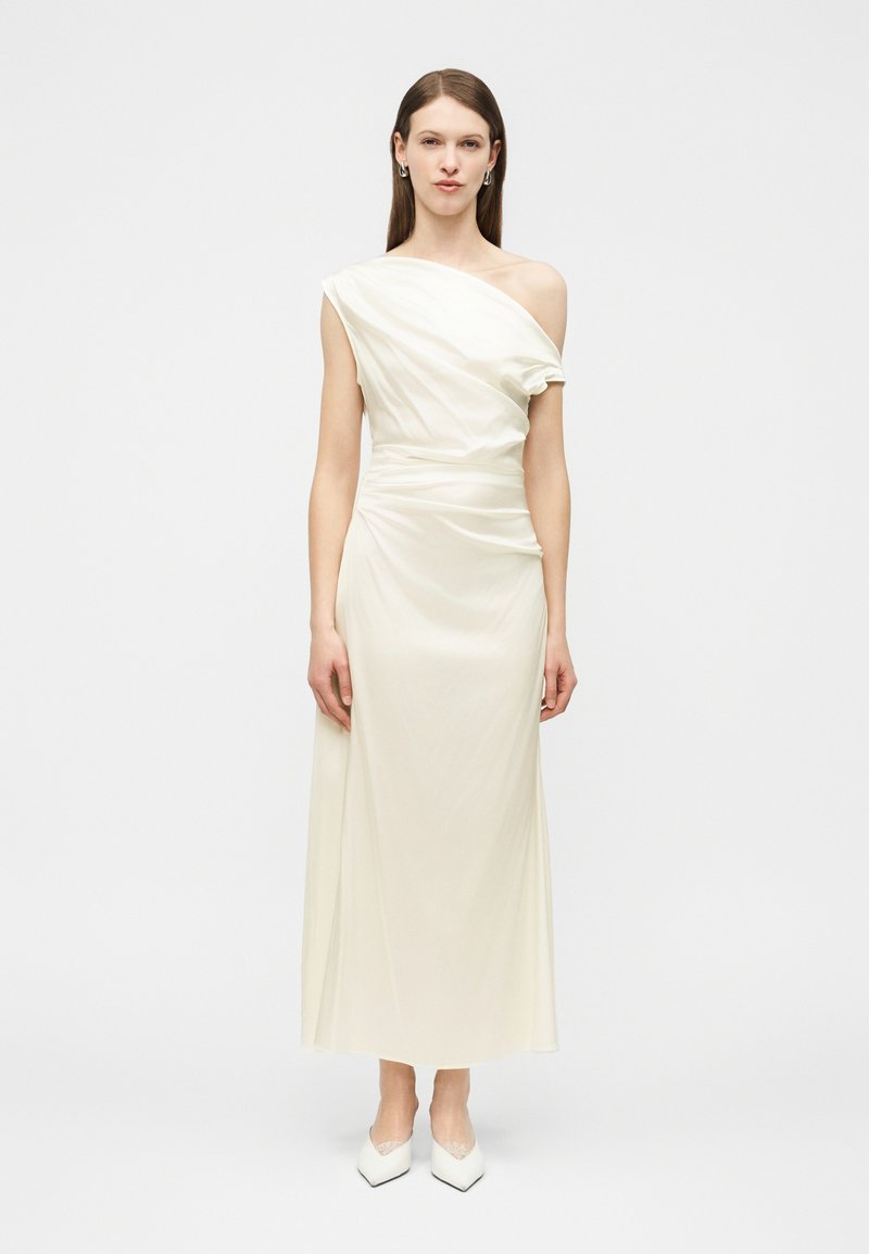 Για γυναίκα που στέκεται, φορόντας μακρύ φόρεμα από σατέν εκρού με έναν ώμο ακάλυπτο και λευκά μυτερά παπούτσια, μπροστά σε απλό φόντο.