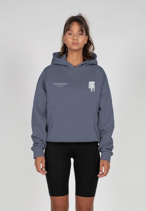 Frau steht frontal, trägt einen blauen Hoodie mit weißem Text und schwarze knielange Shorts vor einem einfarbigen grauen Hintergrund.