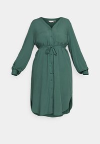 Dlouhý rukáv, košilové šaty v tmavě zelené látce. Má výstřih do V, možné stažení v pase a jednu náprsní kapsu. Hladká textura a délka pod kolena.