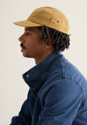 Bărbat cu păr scurt răsucit și cercel mic în formă de cerc, purtând o șapcă bej și o jachetă de denim albastru, privind spre stânga pe un fundal simplu.