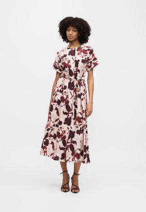 Kobieta o kręconych włosach, ubrana w midi sukienkę z krótkim rękawem i paskiem, z różowo-burgundowym kwiatowym wzorem oraz czarne sandały na obcasie z paskami.