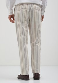 Homme portant un pantalon rayé vertical beige et marron, une chemise blanche et des mocassins marron foncé se tenant devant un fond uni.