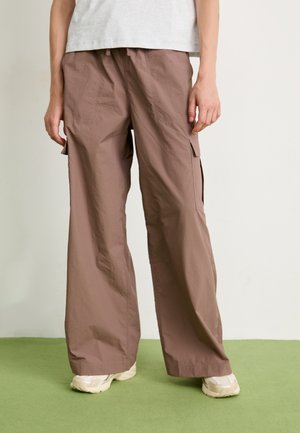 Person som har på seg løse brune cargo-bukser og beige sneakers, står på grønn gulv mot en hvit vegg.