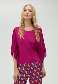Top surdimensionné fuchsia avec une texture douce et des manches larges, assorti à un pantalon à motifs vibrants comportant des formes géométriques en rouge, bleu et beige.