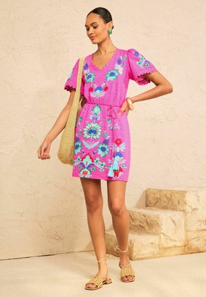 Kobieta w jasnoróżowej, kwiatowo haftowanej sukience z bufkami, niosąca plecioną torbę, stojąca obok kamiennych schodów na beżowym tle.