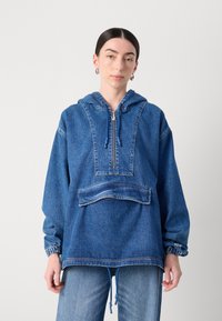 Denimpulloverjacka med huva, fjärdedelsdragkedja, frampocket och elastiska manschetter. Ljus till medelblå färg med en lätt sliten textur.