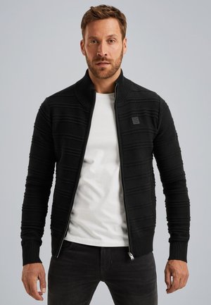 Zwarte ritssluiting vest met een geribbelde textuur en horizontale strepen. Heeft een hoge kraag en is gelaagd over een wit T-shirt.