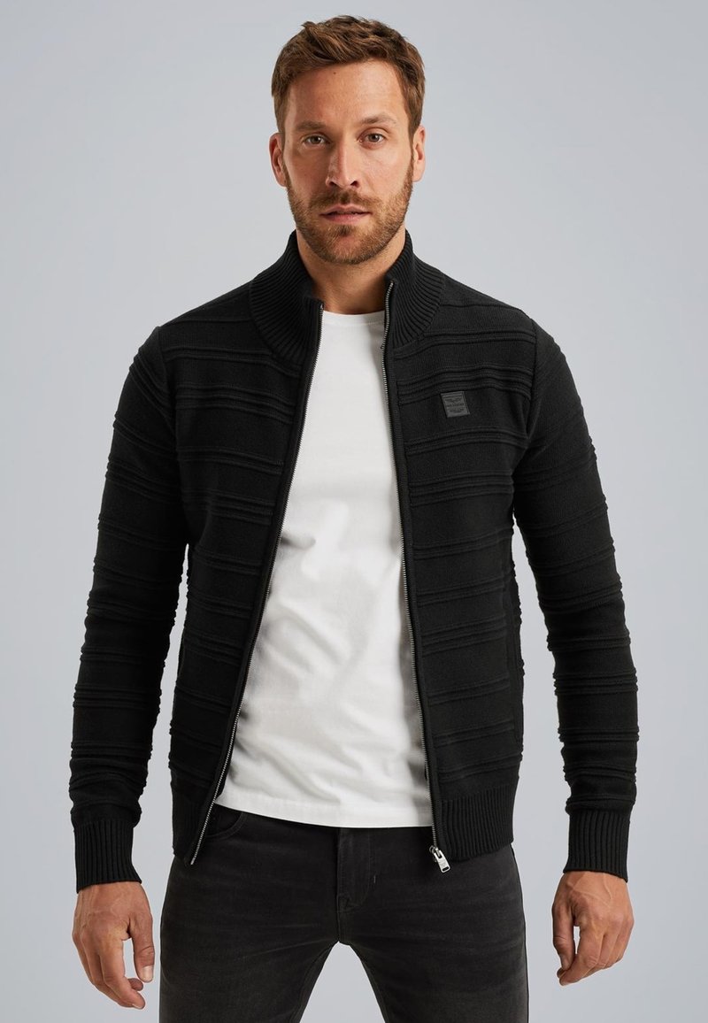 Cardigan zip-up nero con texture a coste e righe orizzontali. Presenta un colletto alto ed è indossato sopra una t-shirt bianca.