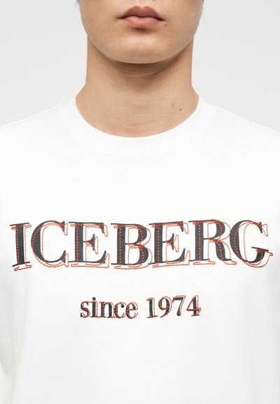 Κοντινό πλάνο ενός ατόμου που φοράει λευκό πουκάμισο με την επιγραφή "ICEBERG" με μεγάλα γράμματα και "since 1974" από κάτω με μικρότερα γράμματα.