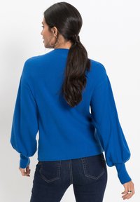 Maglione blu con una texture morbida, maniche lunghe e gonfiate, e orlo aderente. Abbinato a jeans scuri, che mettono in evidenza un design semplice e pulito.