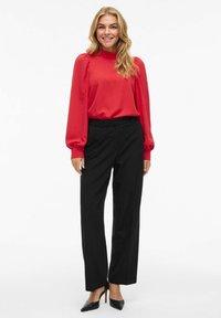 Top rosso a maniche lunghe con arricciature sulle spalle, abbinato a pantaloni neri a vita alta e tacchi a spillo neri. Texture liscia in tutto il capo.