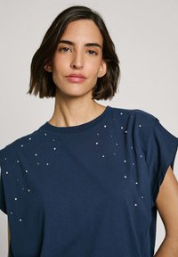 Námořnická modrá trička s krátkým rukávem a kulatým výstřihem, zdobená rozptýlenými kovovými ozdobami pro zvýšení textury a detailu.