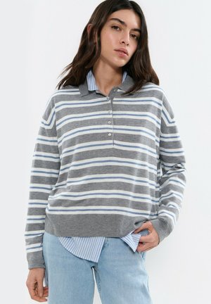 Mujer que lleva un suéter polo de manga larga a rayas grises y blancas sobre una camisa de rayas azules y jeans azul claro, posando con la mano en el bolsillo.