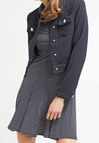 Kapitulerad marinblå jacka med silverknappar över en figurnära svart och vit prickig klänning med en utsvängd kjol och texturerad tyg.