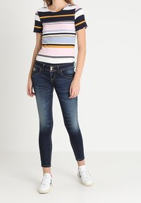 Camiseta de rayas con mangas cortas con volantes en rosa, azul, amarillo y azul marino, combinada con unos jeans oscuros ajustados y zapatillas blancas.